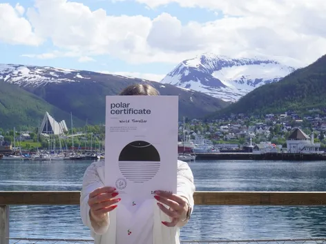 Dame holder opp et Polarsertifikat fra Solivelle om sommeren, med Ishavskatedralen og Tromsdalstinden i bakgrunnen.